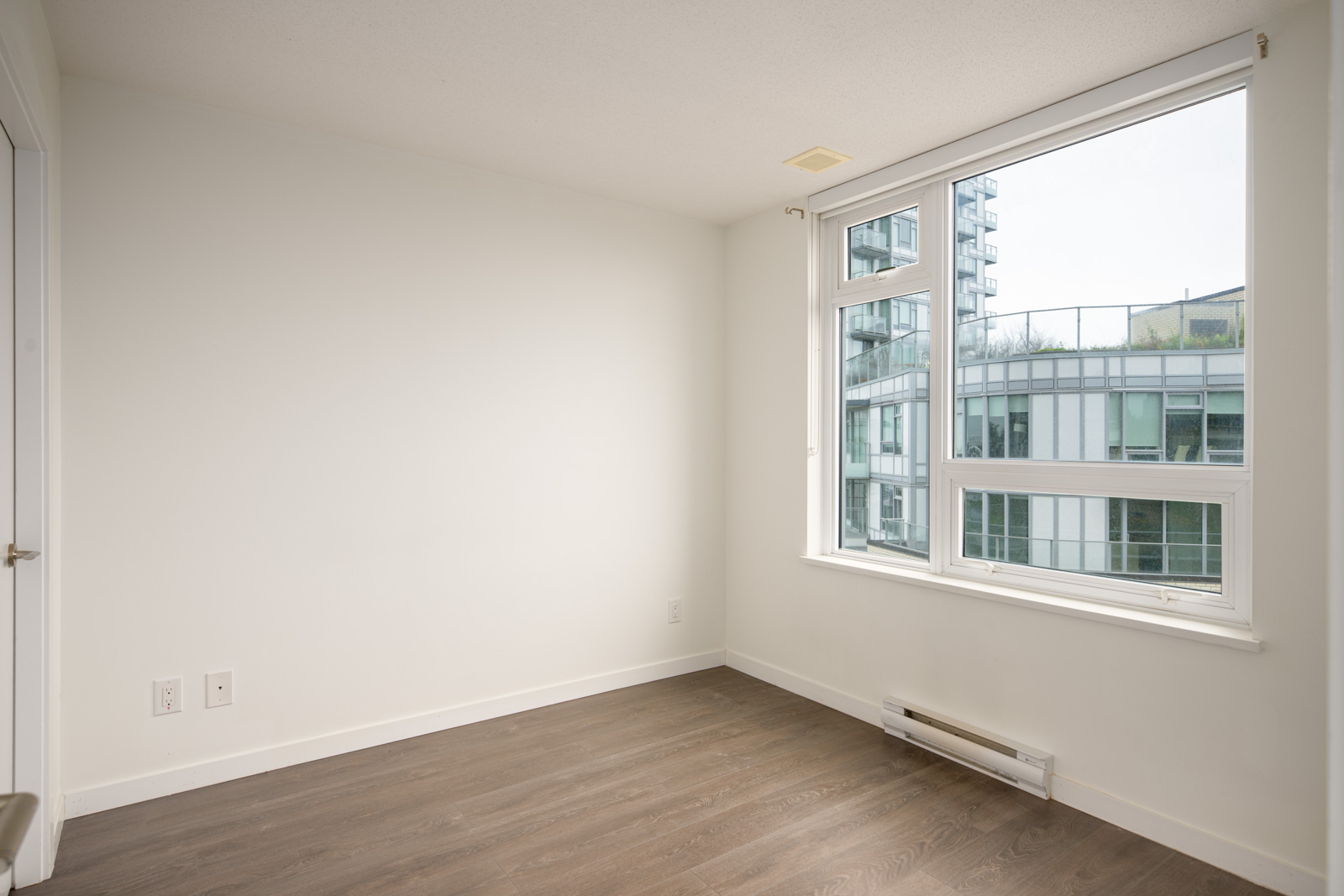 Empty room with white walls, a large window overlooking modern buildings, wood-style flooring, and baseboard heater under the window.