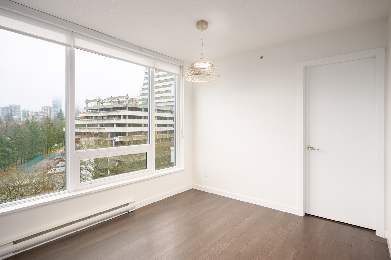 A bright, empty room with white walls, dark hardwood floors, a hanging ceiling light, large windows, and a closed white door. City buildings and trees are visible outside.