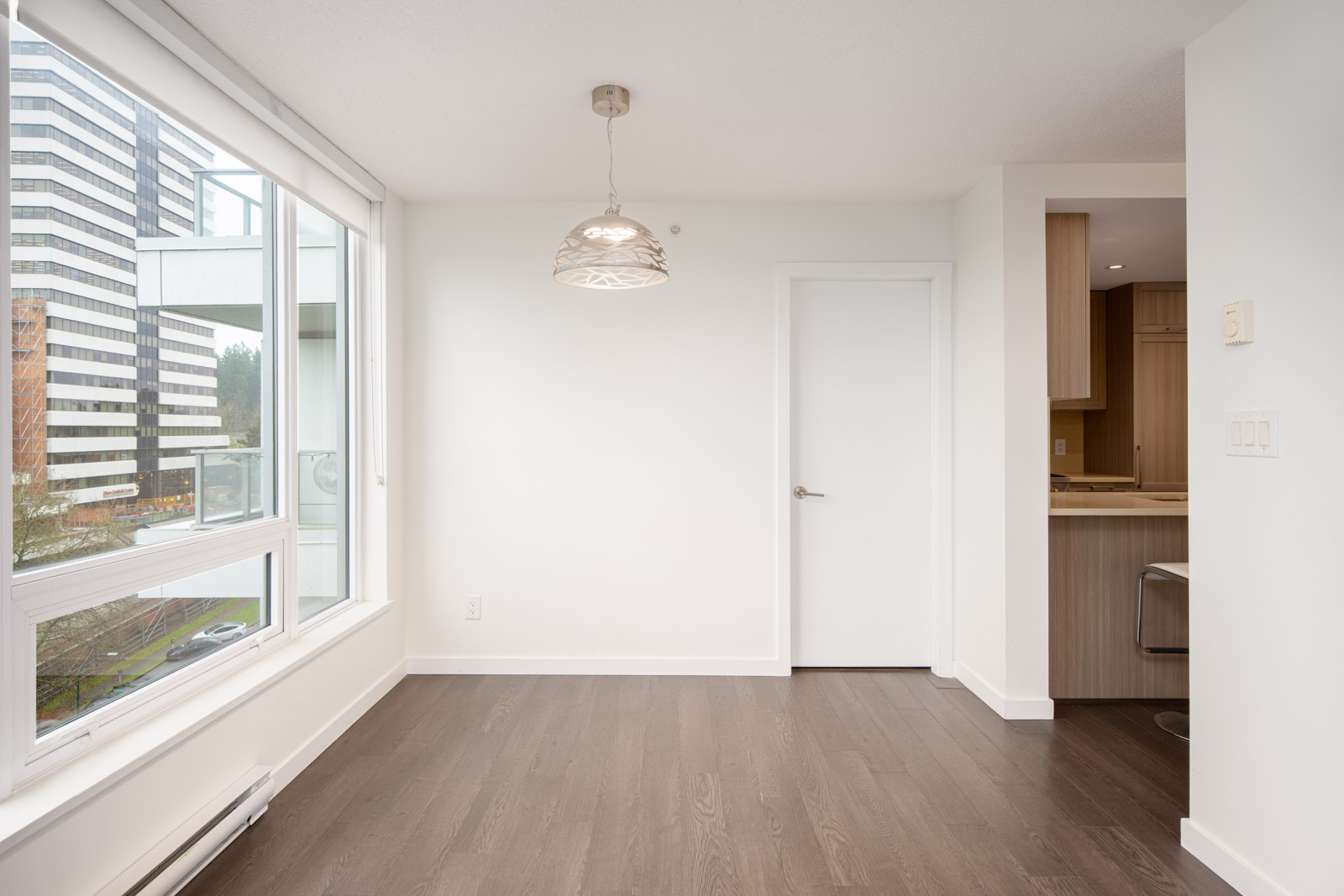 Empty modern room with dark wood flooring, white walls, a window, a hanging light fixture, and an open doorway leading to a kitchen area.