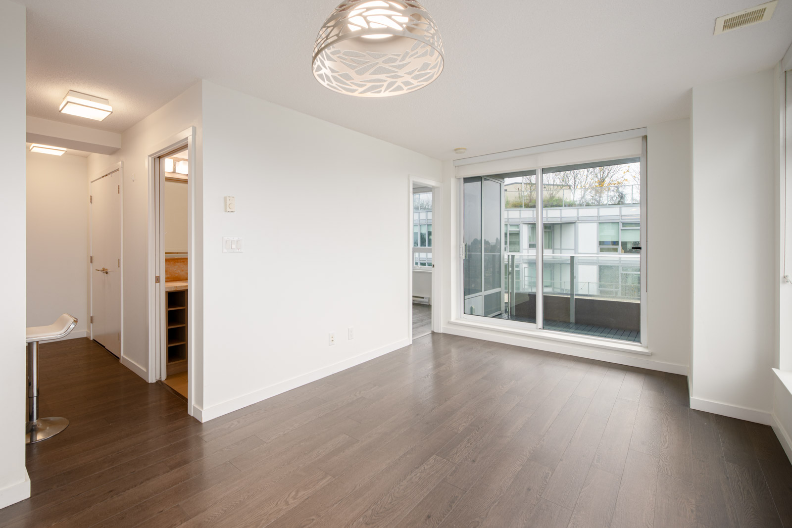 Unfurnished modern apartment with dark wood floors, white walls, large window, glass balcony door, and built-in lighting fixture on the ceiling.