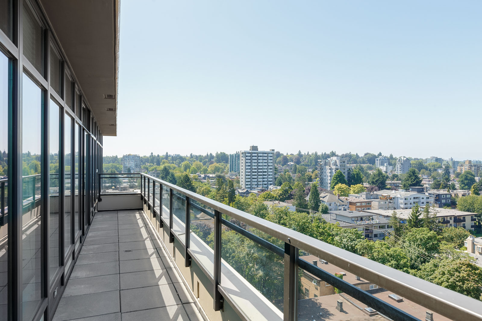A long balcony with glass railing overlooks a cityscape with mid-rise buildings, trees, and a clear blue sky.
