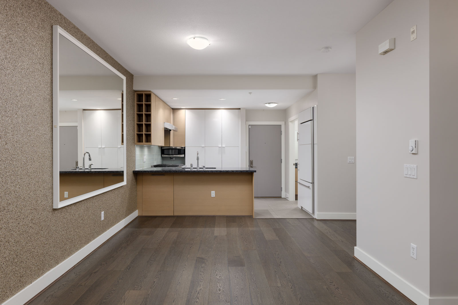 Modern empty apartment with dark wood floors, a large wall mirror, and an open kitchen featuring white cabinets and stainless steel appliances.