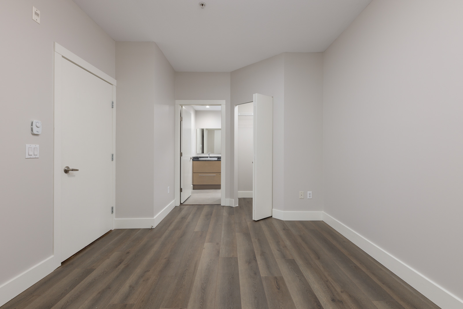 Empty room with light-colored walls, wood flooring, a closed door on the left, and an open doorway leading to a bathroom with a vanity and mirror.