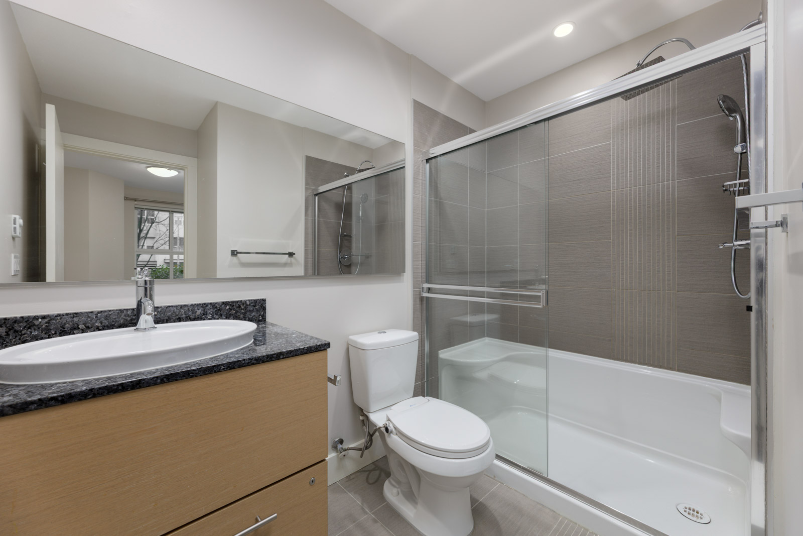 Modern bathroom with a wooden vanity, white sink, toilet, and a bathtub with sliding glass doors and tiled walls. Large mirror above the sink reflects part of the room.
