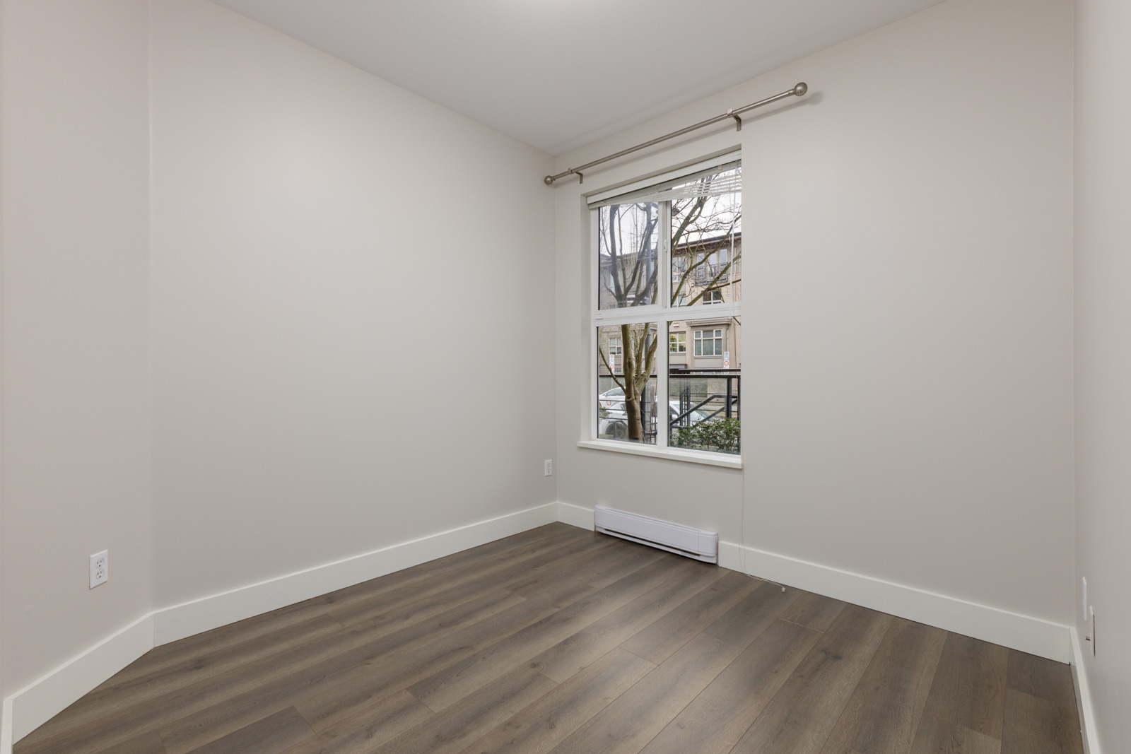 Empty room with light-colored walls, wood flooring, a window with a metal curtain rod, and a baseboard heater below the window.