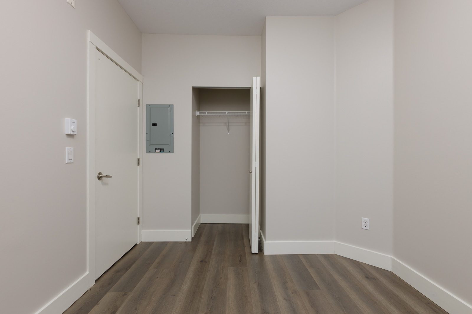 An empty room with light-colored walls, wood flooring, a white door, a small closet with an open door, and an electrical panel on the wall.