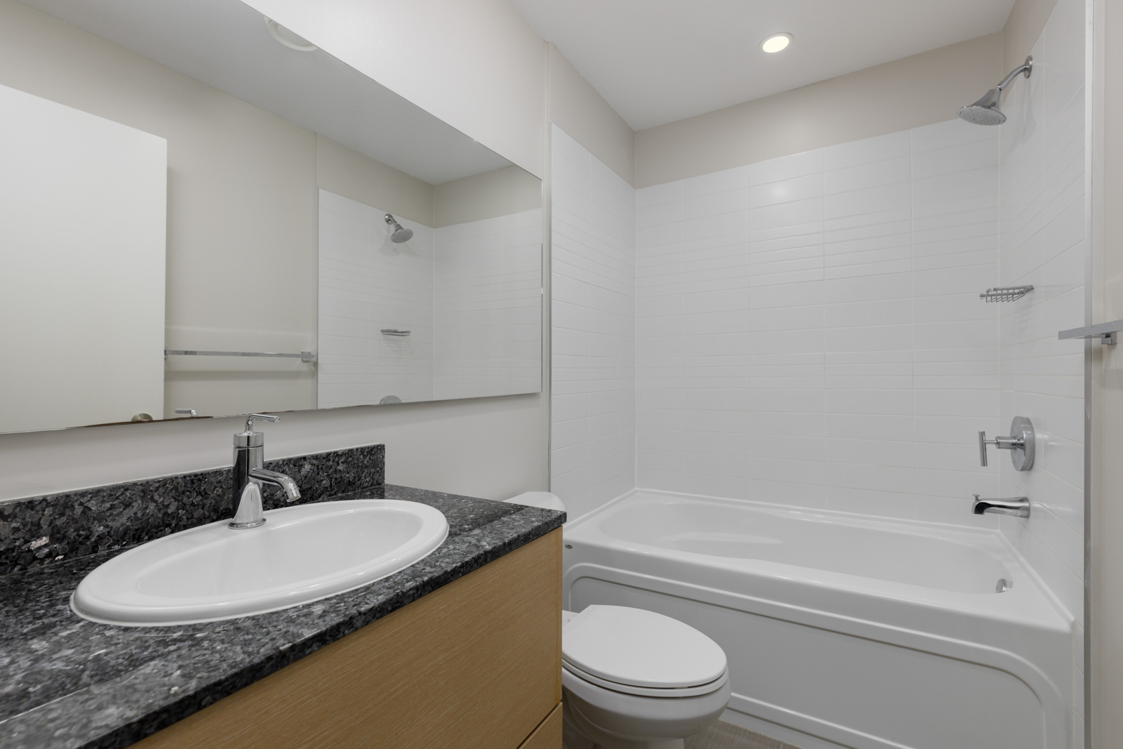 Modern bathroom with a granite countertop sink, wooden vanity, toilet, and a white bathtub-shower combination with tiled walls.