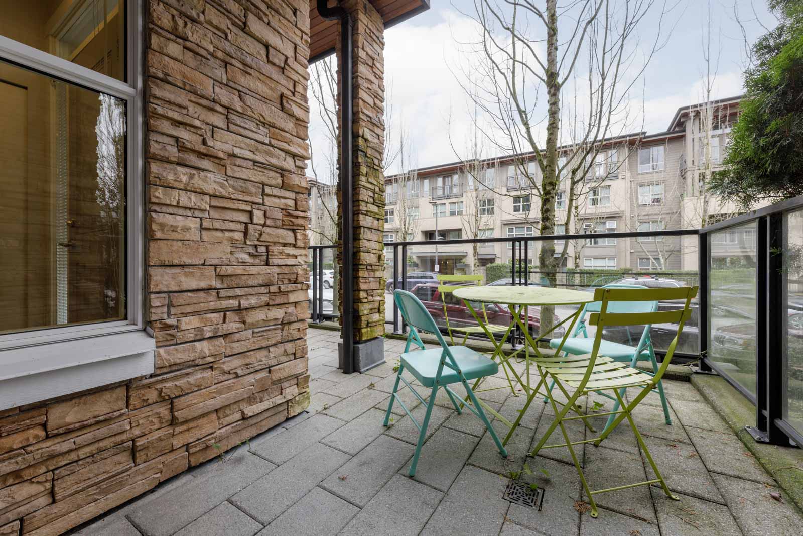 Small patio with a round green metal table and three mismatched chairs, surrounded by a glass railing and adjacent to a stone wall, overlooking a residential complex.