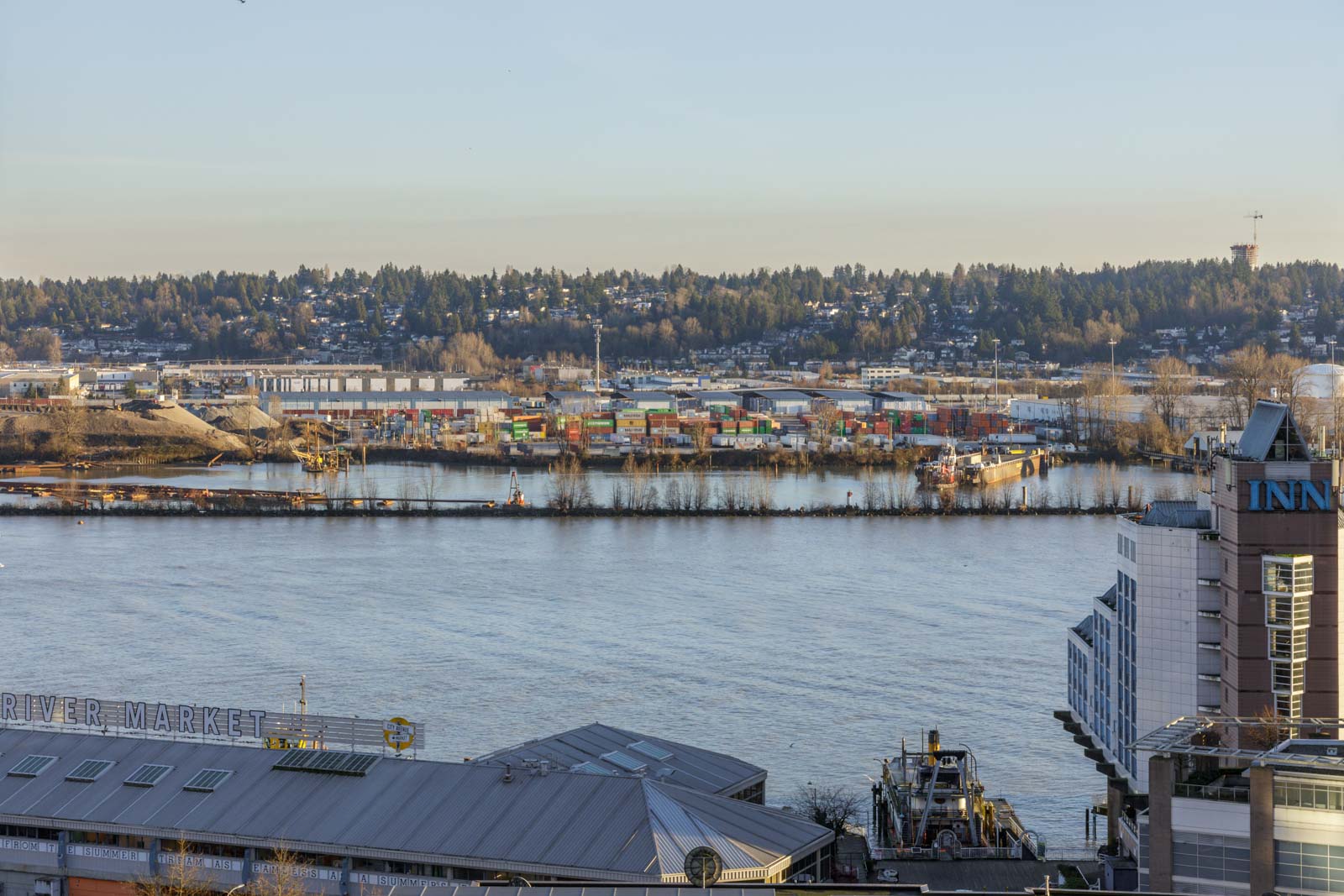 A river with a waterfront market building in the foreground, colorful shipping containers and industrial areas across the water, and a tree-covered hillside in the background.