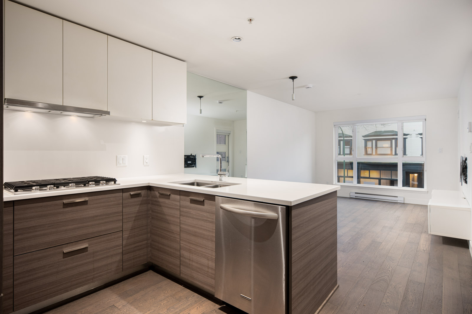 Modern kitchen with wood cabinets, stainless steel appliances, and white countertops, open to a living area with large window and dark wood flooring.