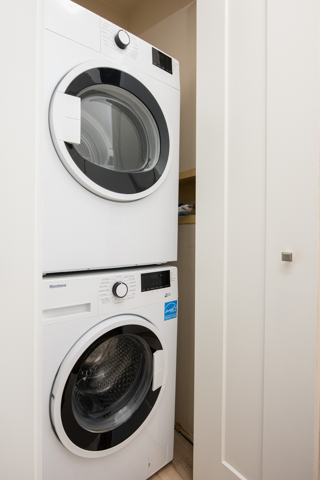 Stacked front-loading washer and dryer in a small laundry closet with white walls and a partially open door.