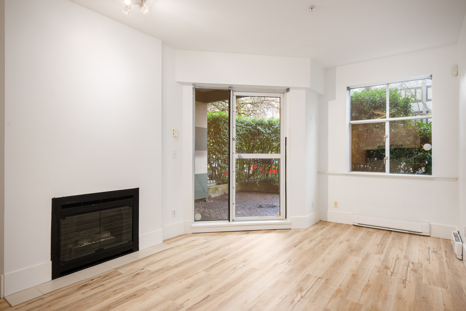 Bright, empty living room with light wood flooring, a fireplace on the left, large sliding doors leading to a patio, and a window on the right overlooking greenery.