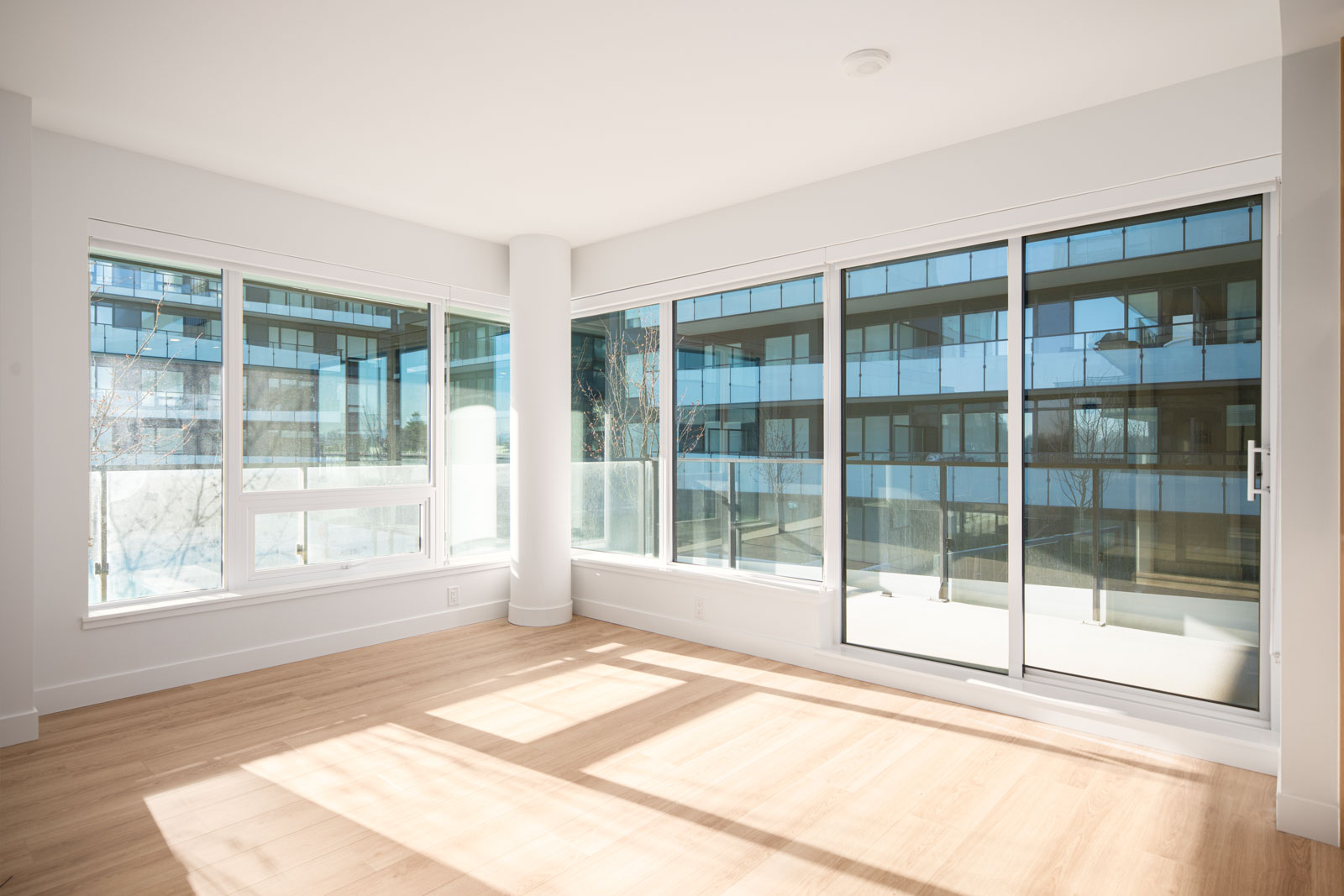 Bright, empty room with large floor-to-ceiling windows, light wood flooring, and a view of a modern apartment building exterior.