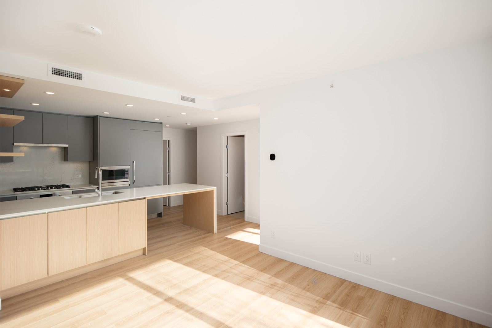 Modern, unfurnished kitchen and living area with light wood flooring, white walls, grey cabinets, built-in appliances, and a large counter island. Natural light enters from the right.