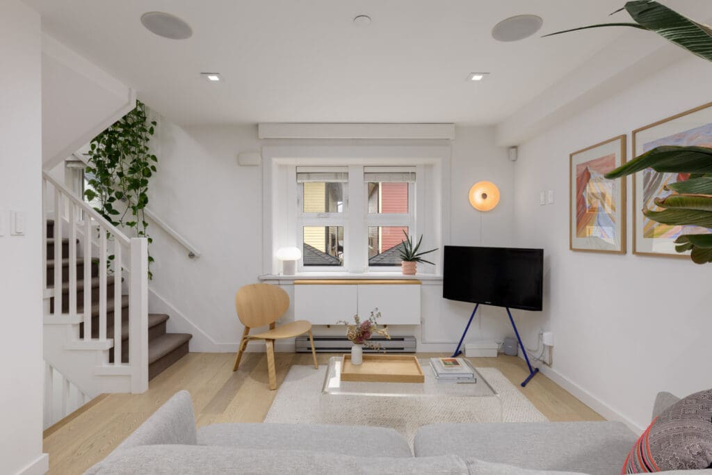 A modern living room with light wood floors, white walls, a small TV on a stand, a wooden chair, framed art, and a staircase with plants.