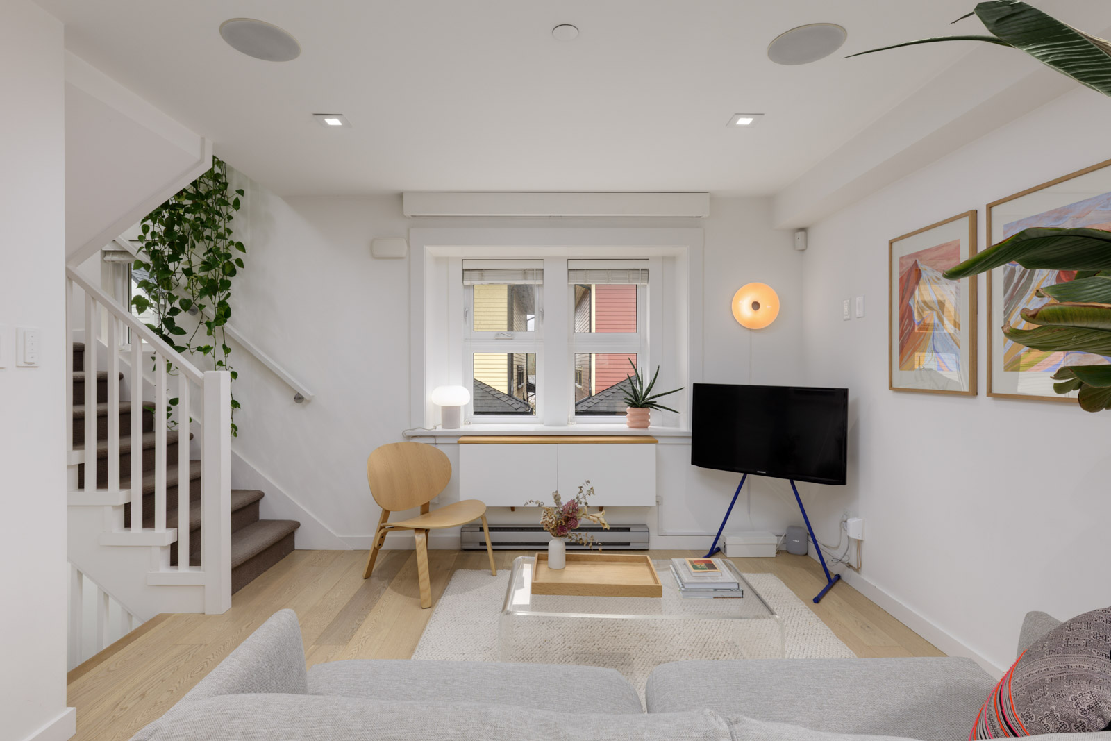 A modern living room with light wood floors, white walls, a small TV on a stand, a wooden chair, framed art, and a staircase with plants.