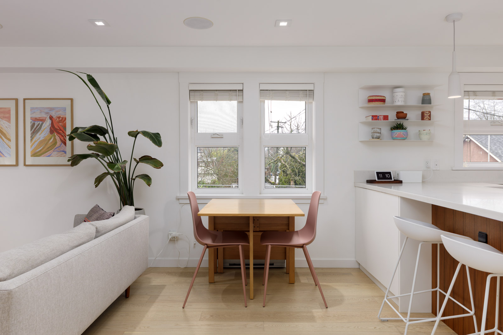 A modern dining area with a wooden table, two pink chairs, and nearby kitchen counter with two white barstools, set in a bright room with large windows and minimal decor.