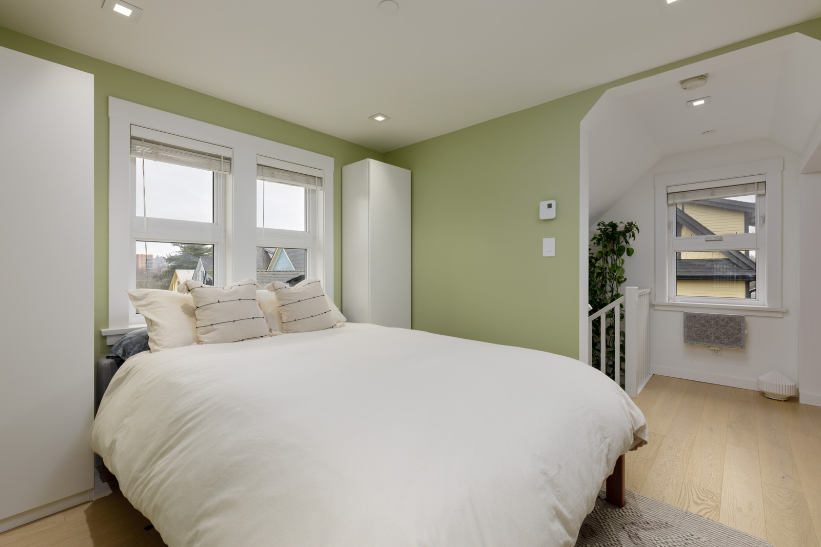 A bedroom with light green walls, a white bed with pillows, two windows, wood flooring, and an open doorway leading to a stairway.