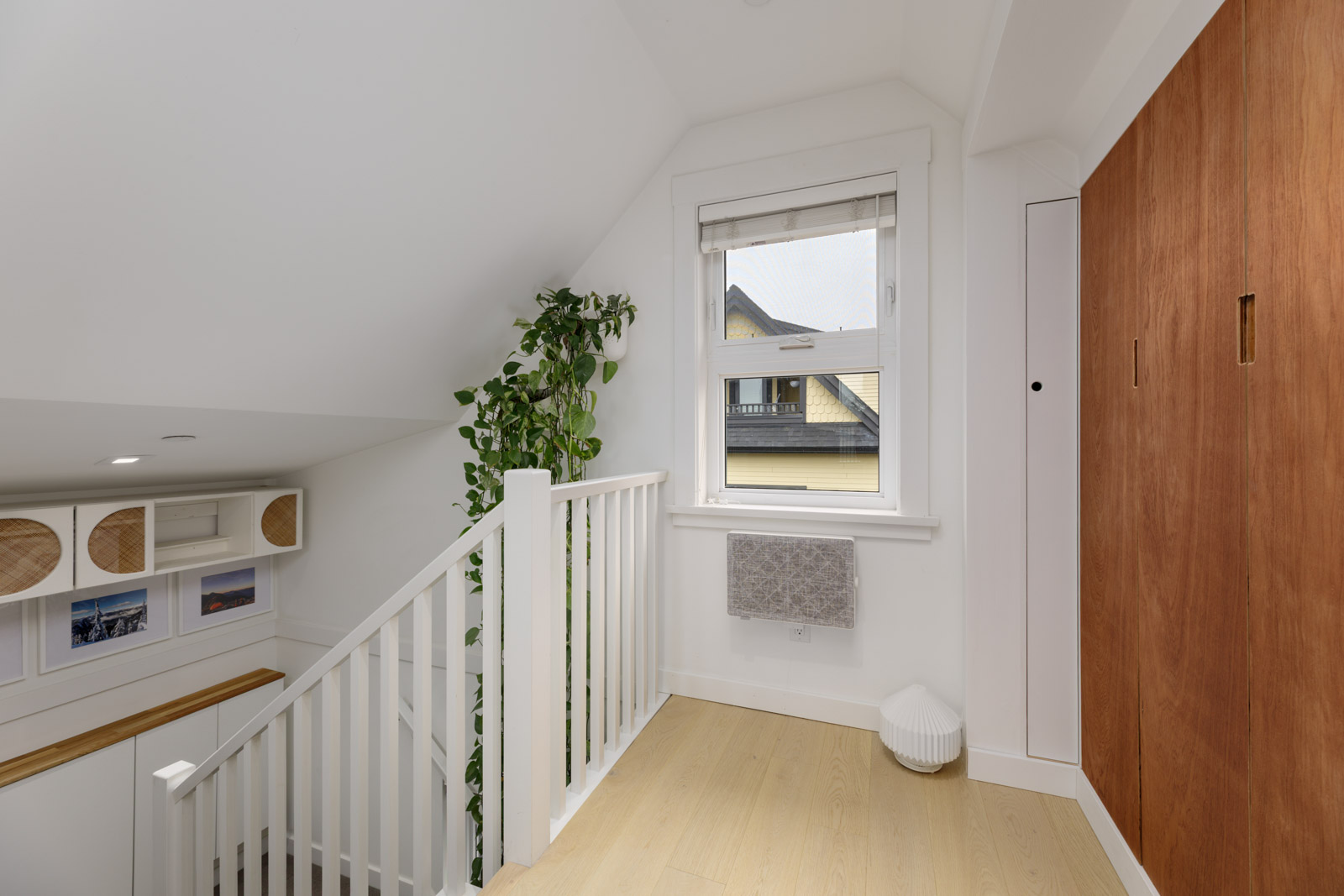A bright upstairs landing with a window, wooden flooring, a white railing, a tall green plant, wall art, and wood-paneled closet doors.