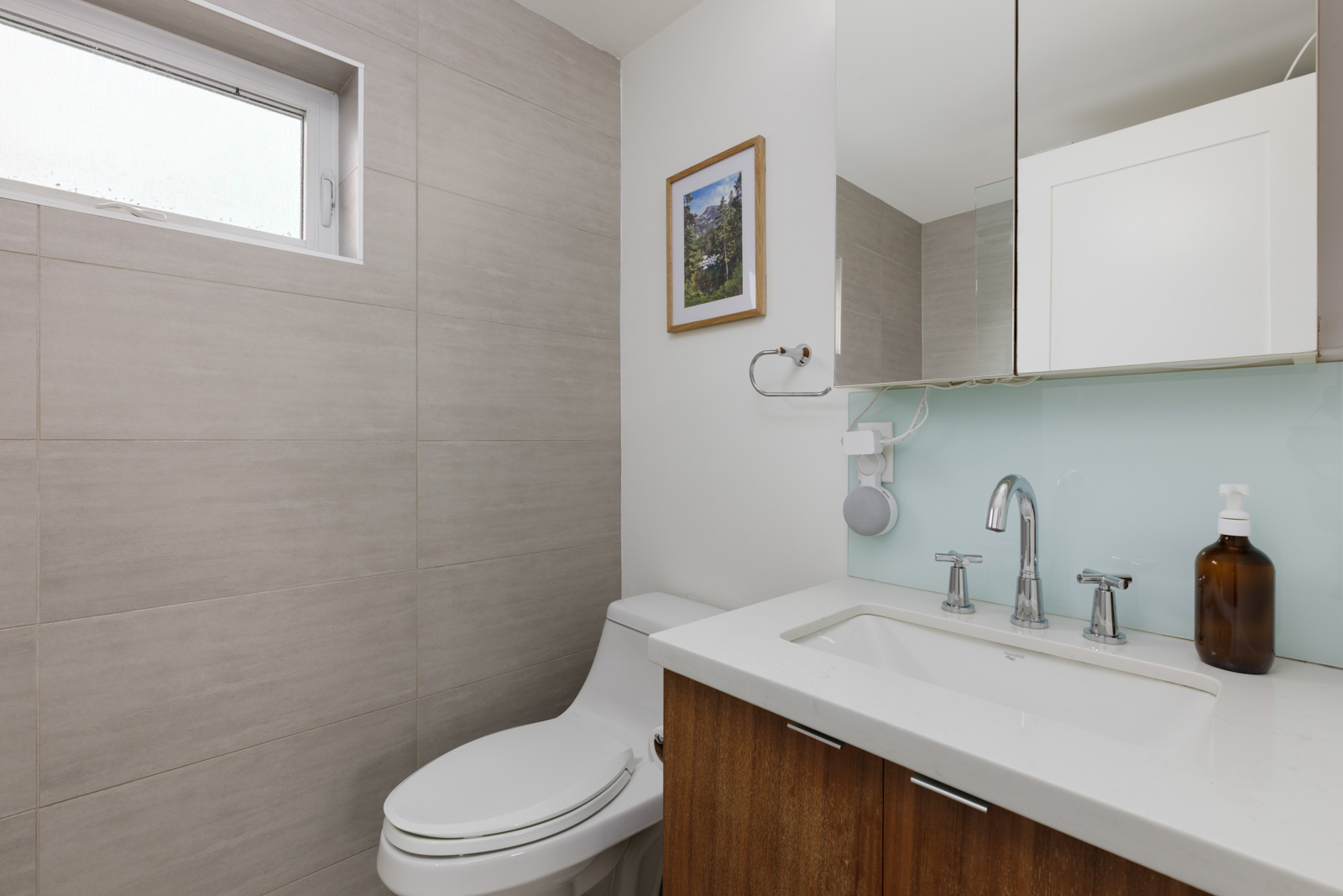 Modern bathroom with a white toilet, wood vanity, white countertop, mounted mirror, wall art, and a soap dispenser, with light gray tiled walls and a small frosted window.
