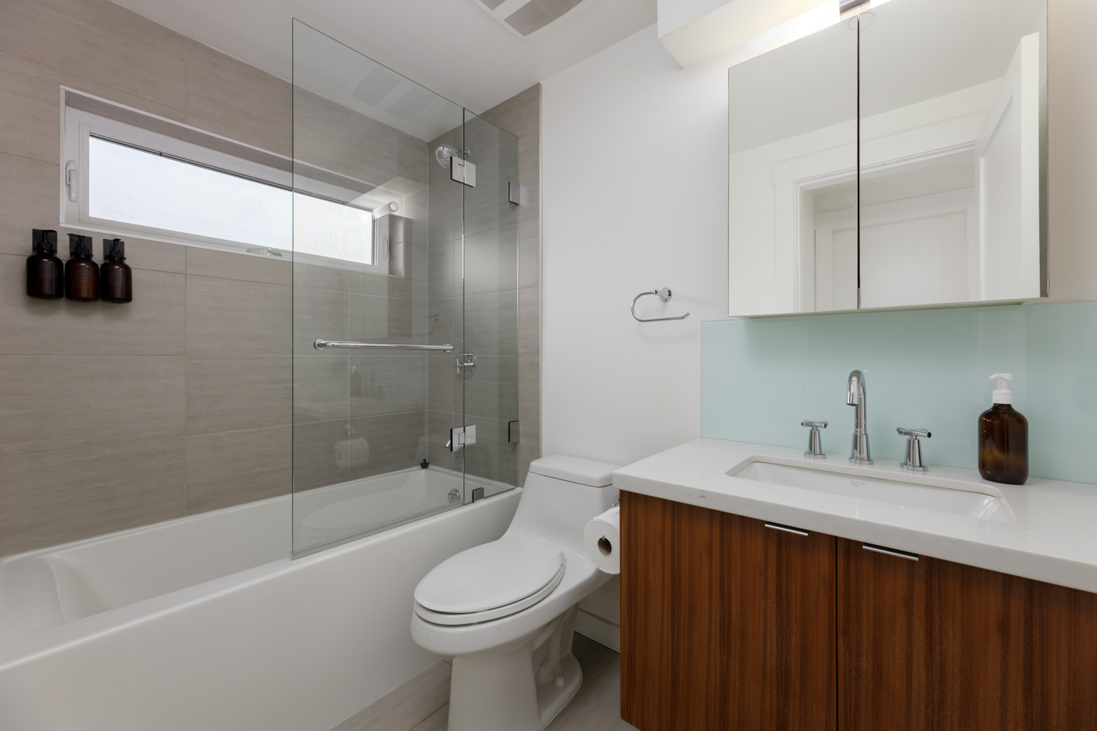 Modern bathroom with a glass-enclosed bathtub, white toilet, wood vanity with sink, wall-mounted faucet, and soap dispensers on the tub wall.