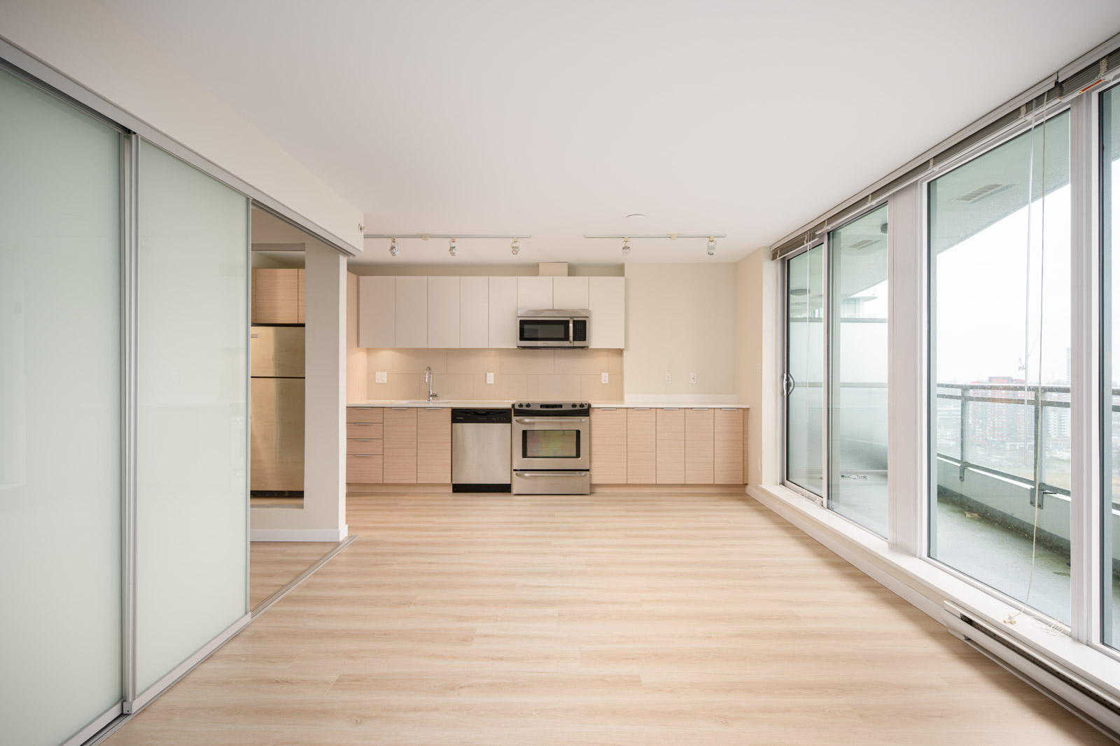 Modern apartment interior with light wood flooring, open kitchen, stainless steel appliances, large windows, sliding glass doors, and balcony view.