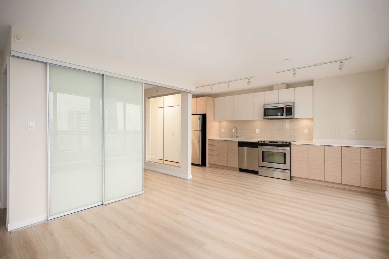 Modern, unfurnished apartment with light wood floors, open kitchen featuring stainless steel appliances, and a room separated by frosted glass sliding doors.