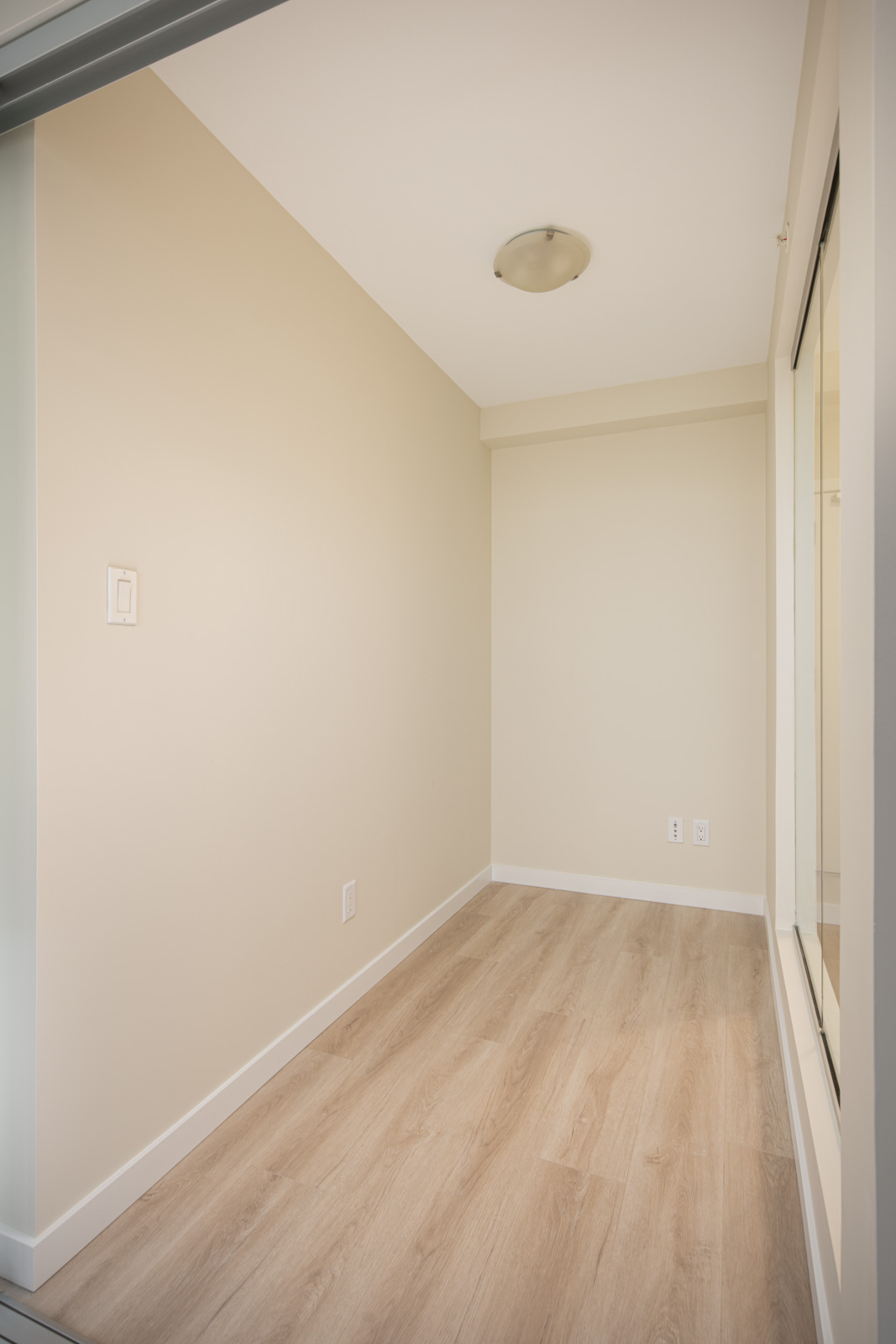 Narrow, empty room with light wood flooring, beige walls, a ceiling light fixture, and a large window on the right side.