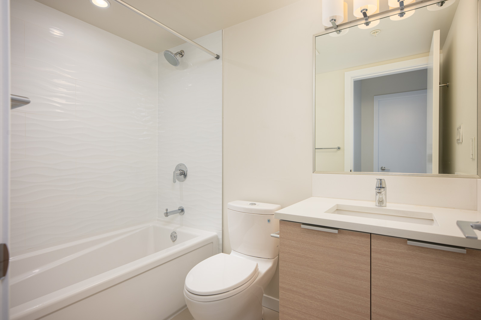 Modern bathroom with a bathtub-shower combo, toilet, light wood vanity with sink, large mirror, and white tile walls.