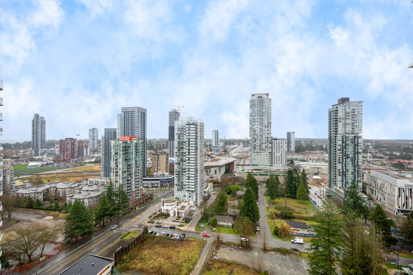 High-rise buildings and construction sites dominate an urban skyline, with some trees and roads visible in the foreground under a partly cloudy sky.