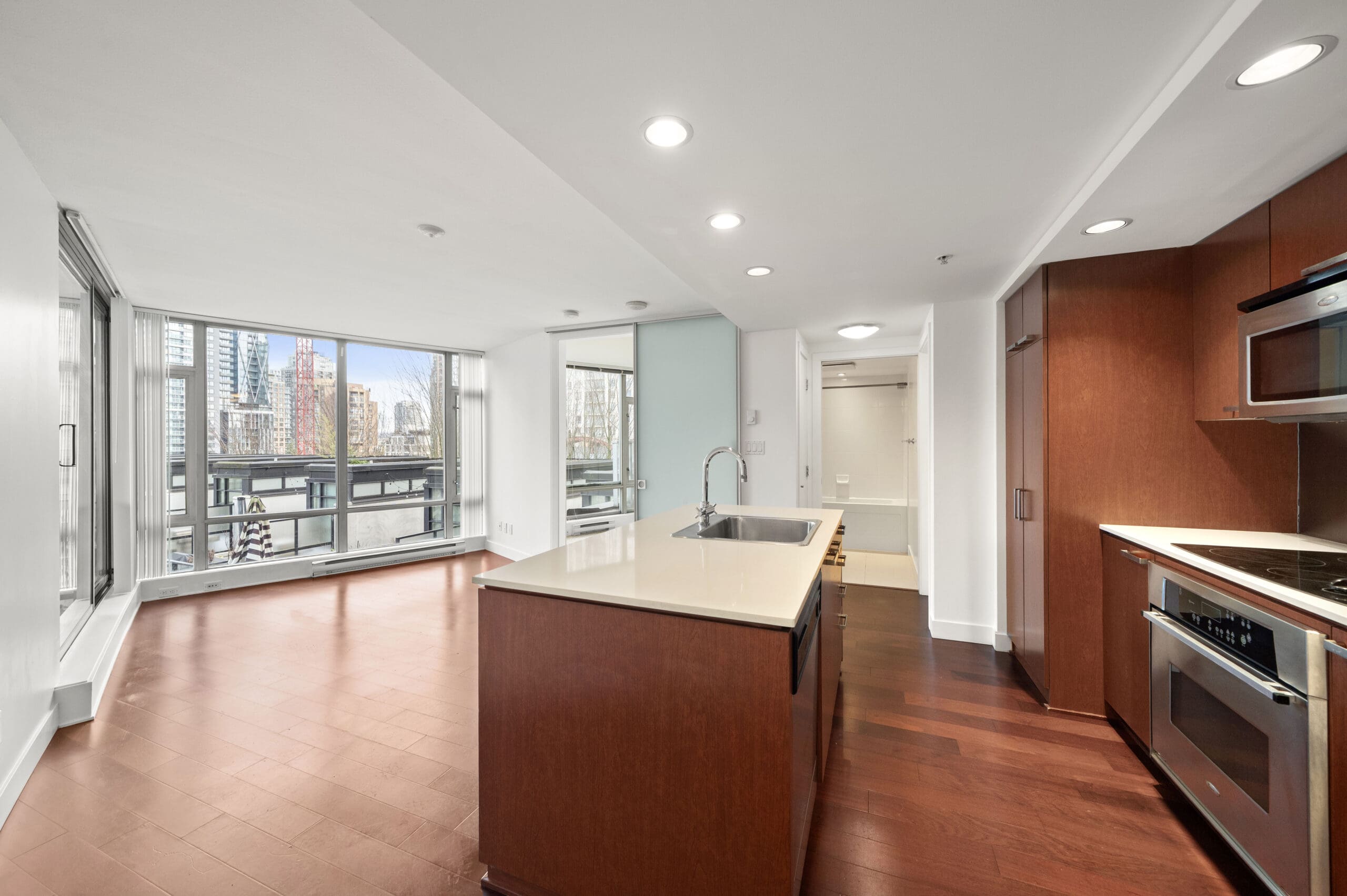 Modern apartment kitchen with an island, stainless steel appliances, and open living area featuring large windows overlooking a cityscape.