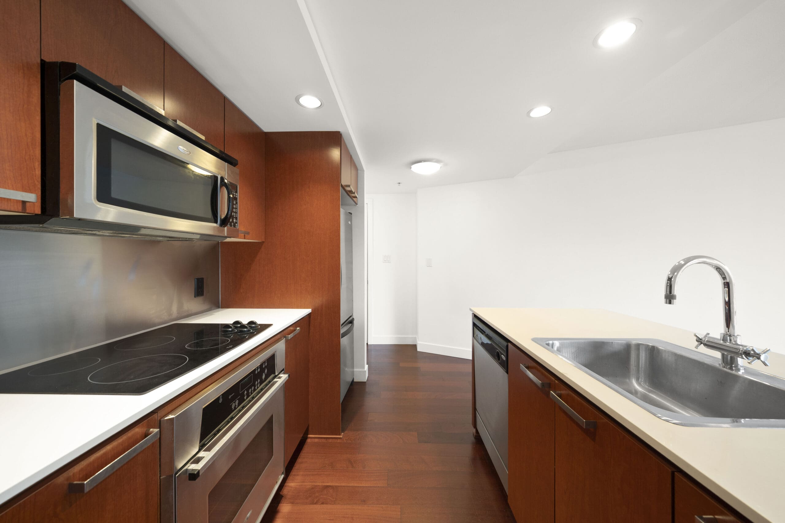 Modern kitchen with wooden cabinets, stainless steel appliances, electric stove, built-in microwave, sink, and dishwasher, with white walls and dark hardwood floors.