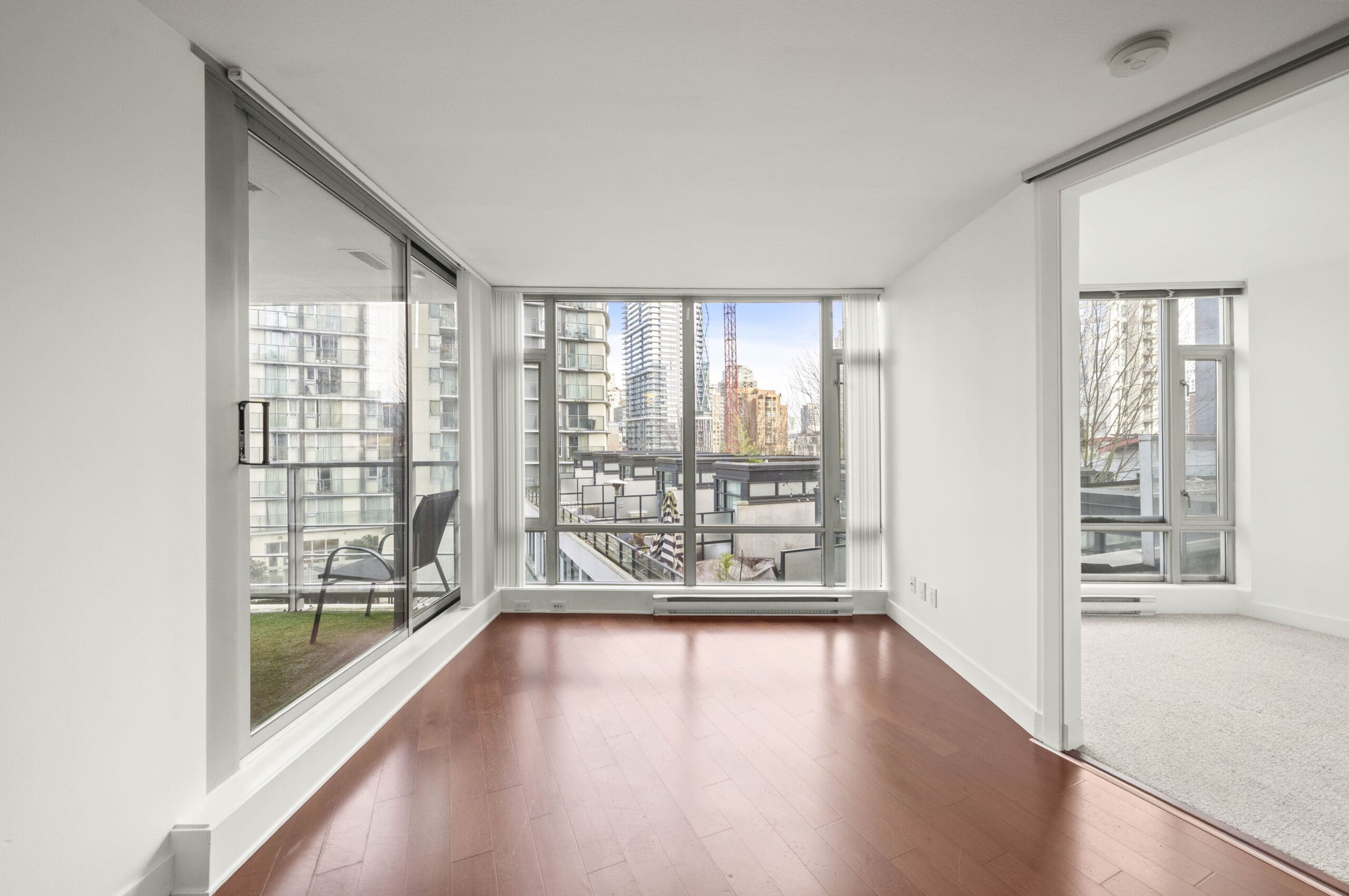 Bright modern apartment with large windows, hardwood floors, glass balcony, and a view of city buildings; adjacent room visible through an open sliding door.