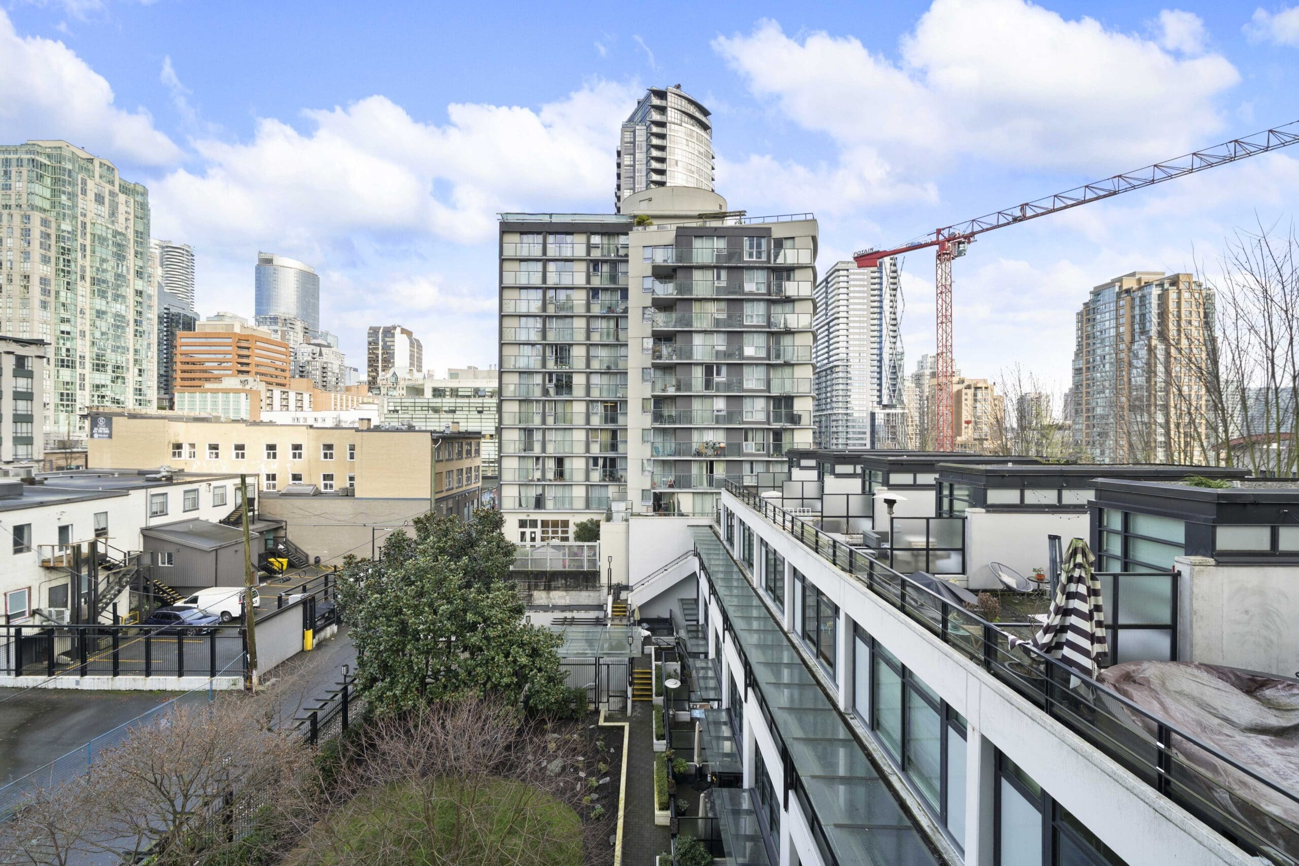 View of modern city buildings with glass windows, a construction crane, and a small green courtyard in the foreground under a partly cloudy sky.