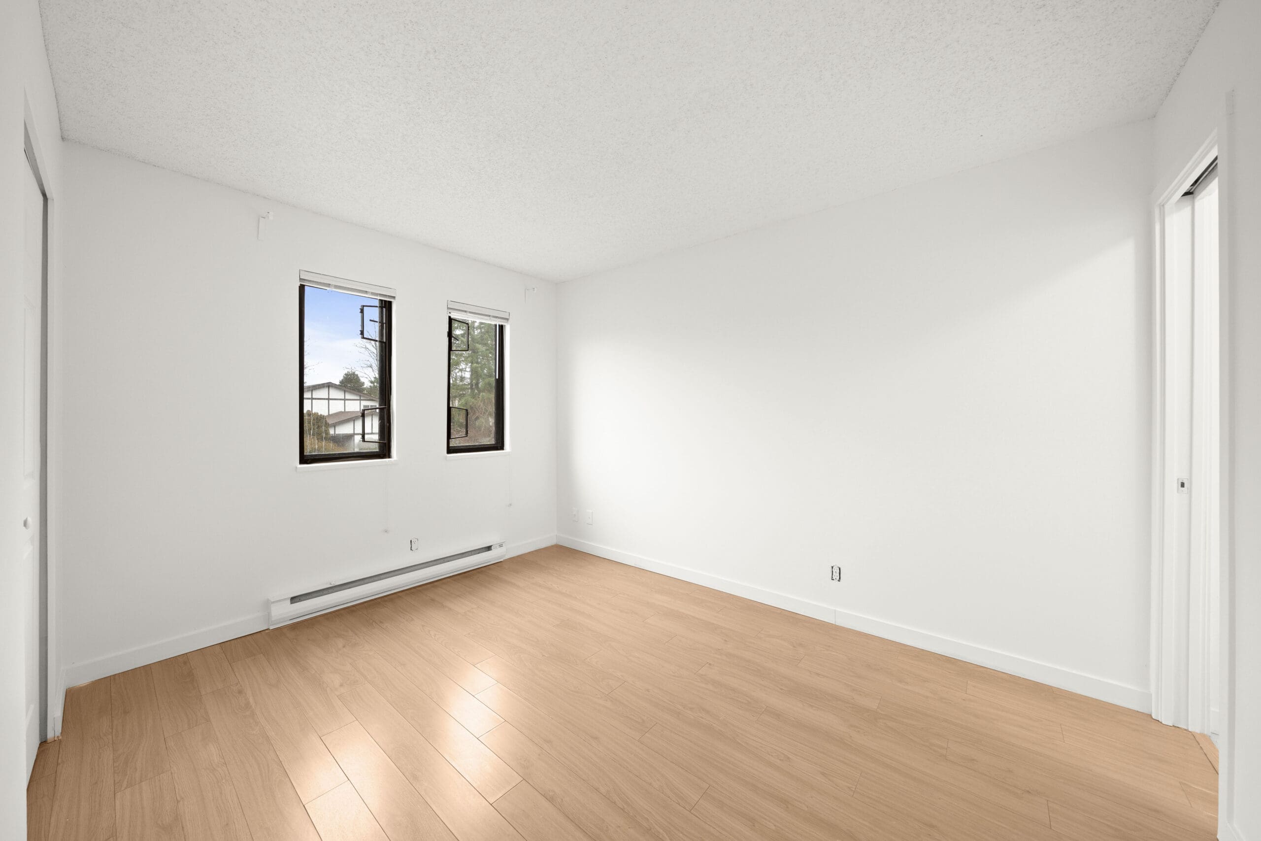 Empty room with light wood flooring, white walls, two windows, baseboard heater, and double closets on opposite sides.