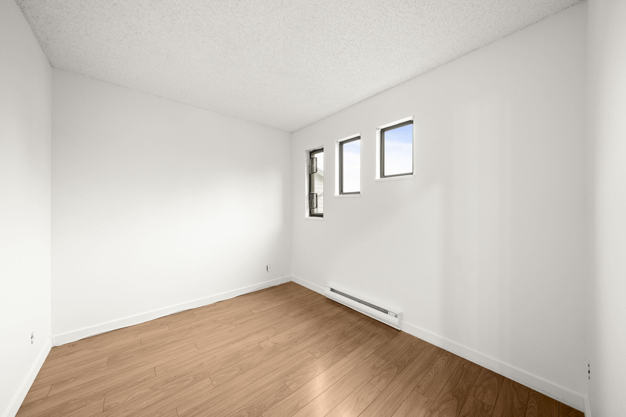 Empty room with white walls, light wood flooring, and three small rectangular windows letting in natural light.