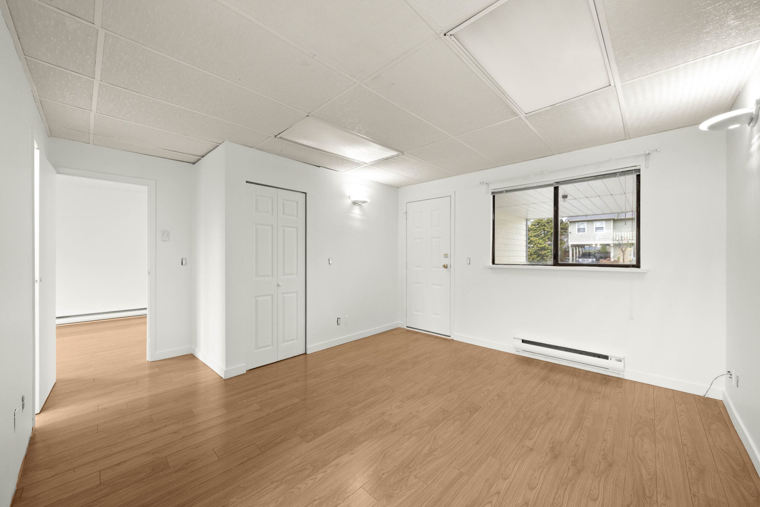 Empty room with white walls, wood flooring, a window with a view outside, built-in closet, closed door, and ceiling panel lights.