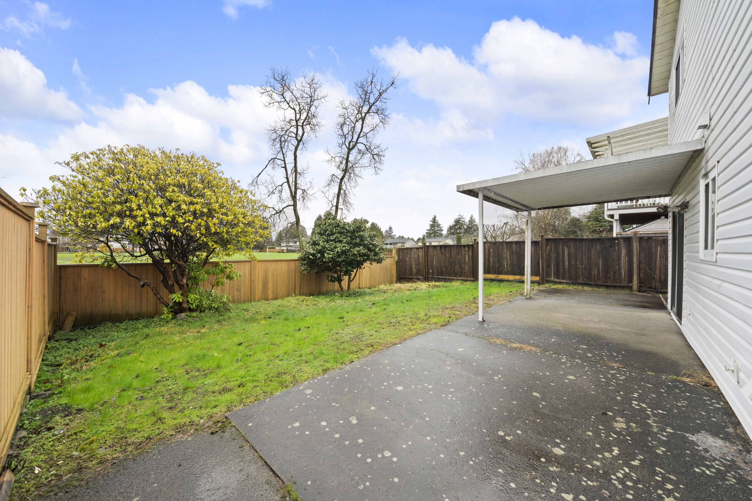 A backyard with a concrete patio, covered area, grassy lawn, a few trees and shrubs, and wooden fencing under a partly cloudy sky.