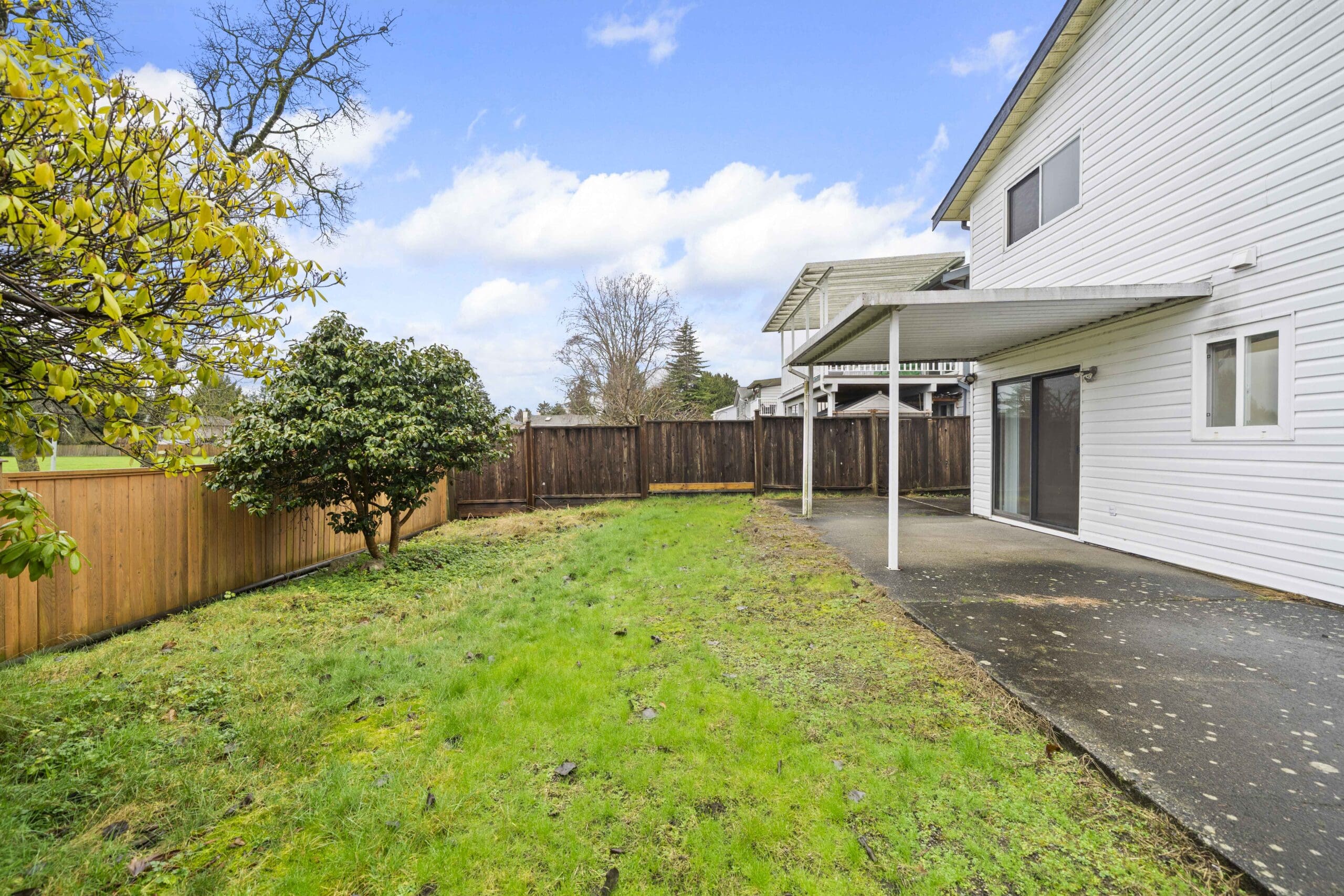 A backyard with a patchy grass lawn, a tree, a wooden fence, and a white house featuring a covered concrete patio and sliding glass doors.