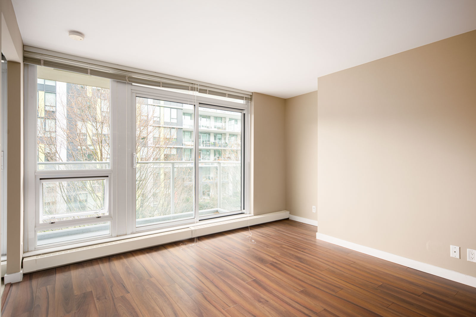 Empty room with beige walls, large floor-to-ceiling windows, wood laminate flooring, and a view of modern apartment buildings outside.