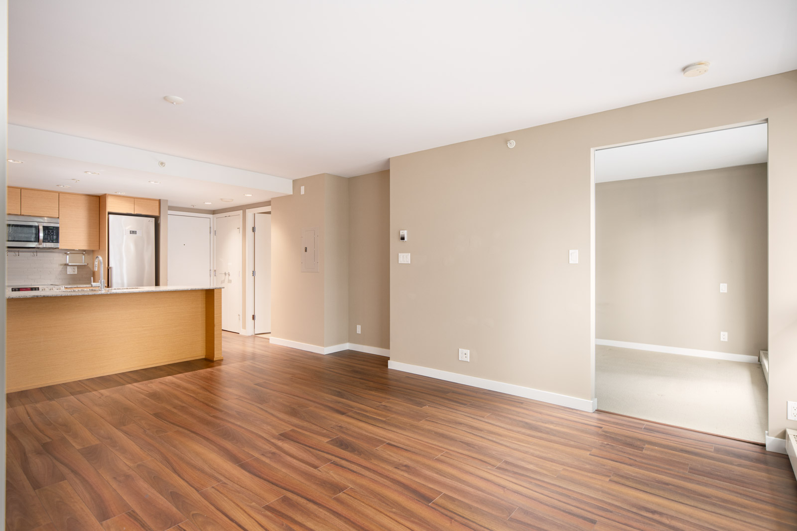 Unfurnished apartment with wood-look flooring, beige walls, open kitchen area with stainless appliances, and an adjacent empty room with carpet.