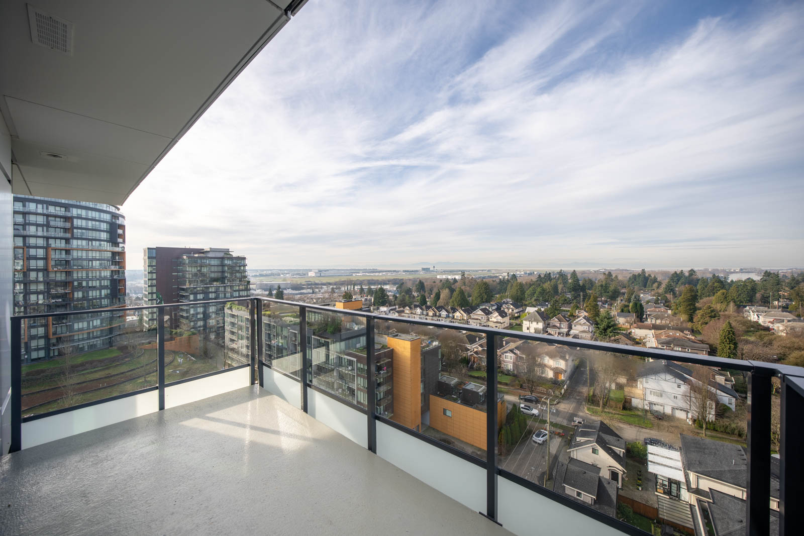 View from a high-rise balcony showing glass railings, nearby buildings, residential streets, and a partly cloudy sky over a suburban neighborhood.