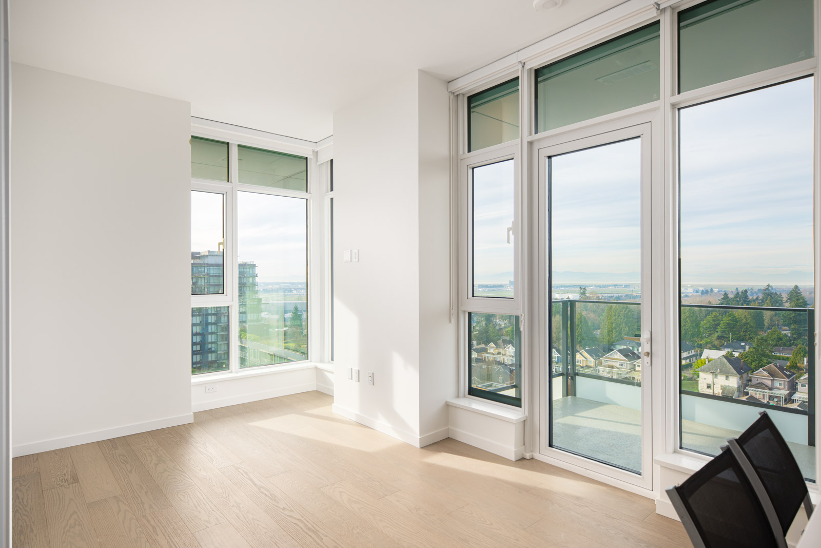 Bright modern apartment interior with large windows, light wood floors, and a view of city buildings and greenery outside on a sunny day.