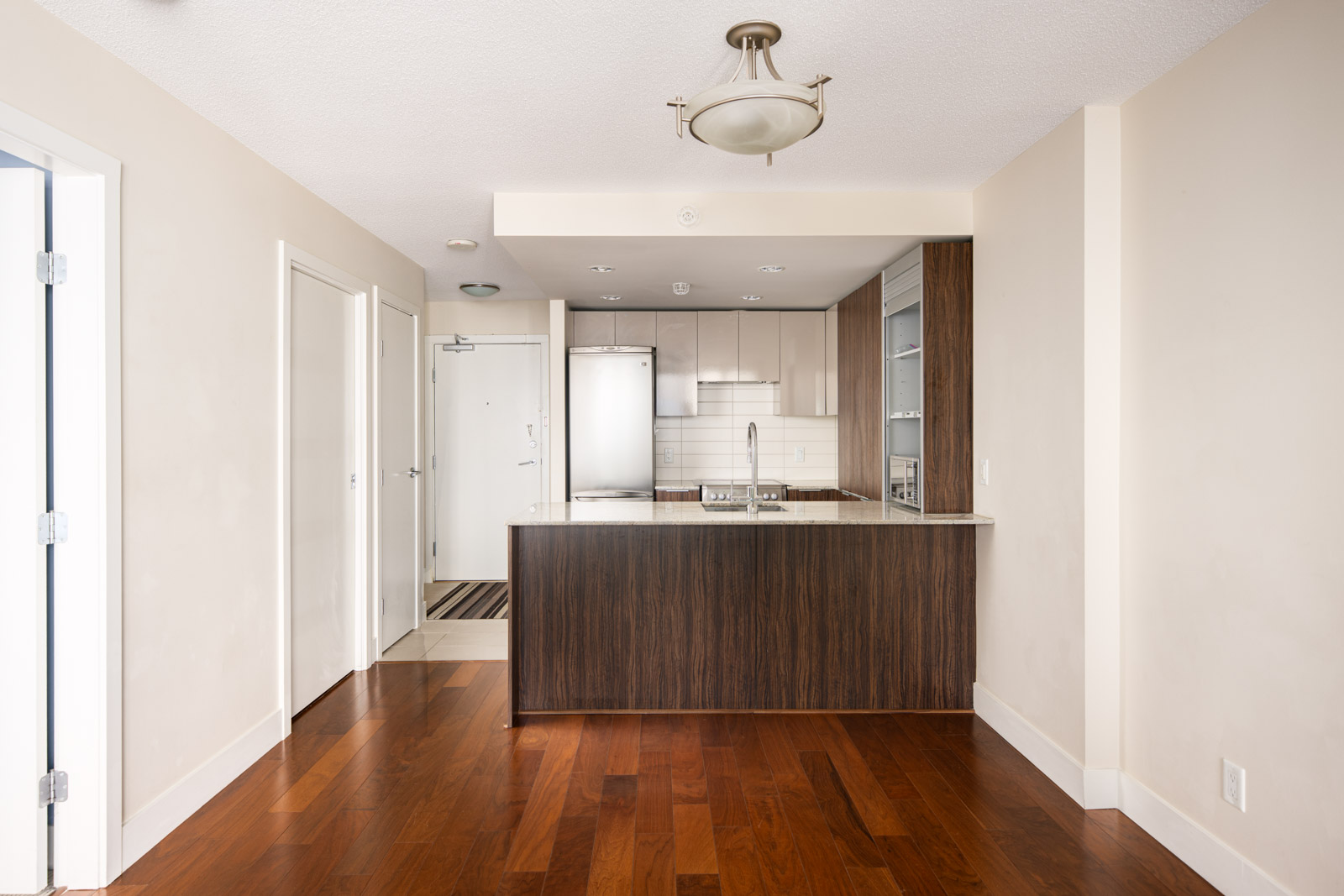 Modern kitchen with wood floors, a dark wood island, white countertops, stainless steel appliances, and white walls. A ceiling light fixture hangs above the kitchen area.