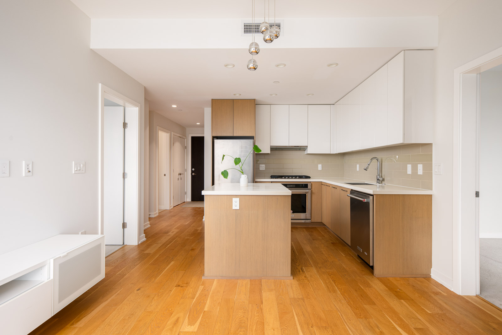Modern kitchen with light wood floors, white walls, an island, stainless steel appliances, and minimalist cabinetry; doors and hallway visible in the background.