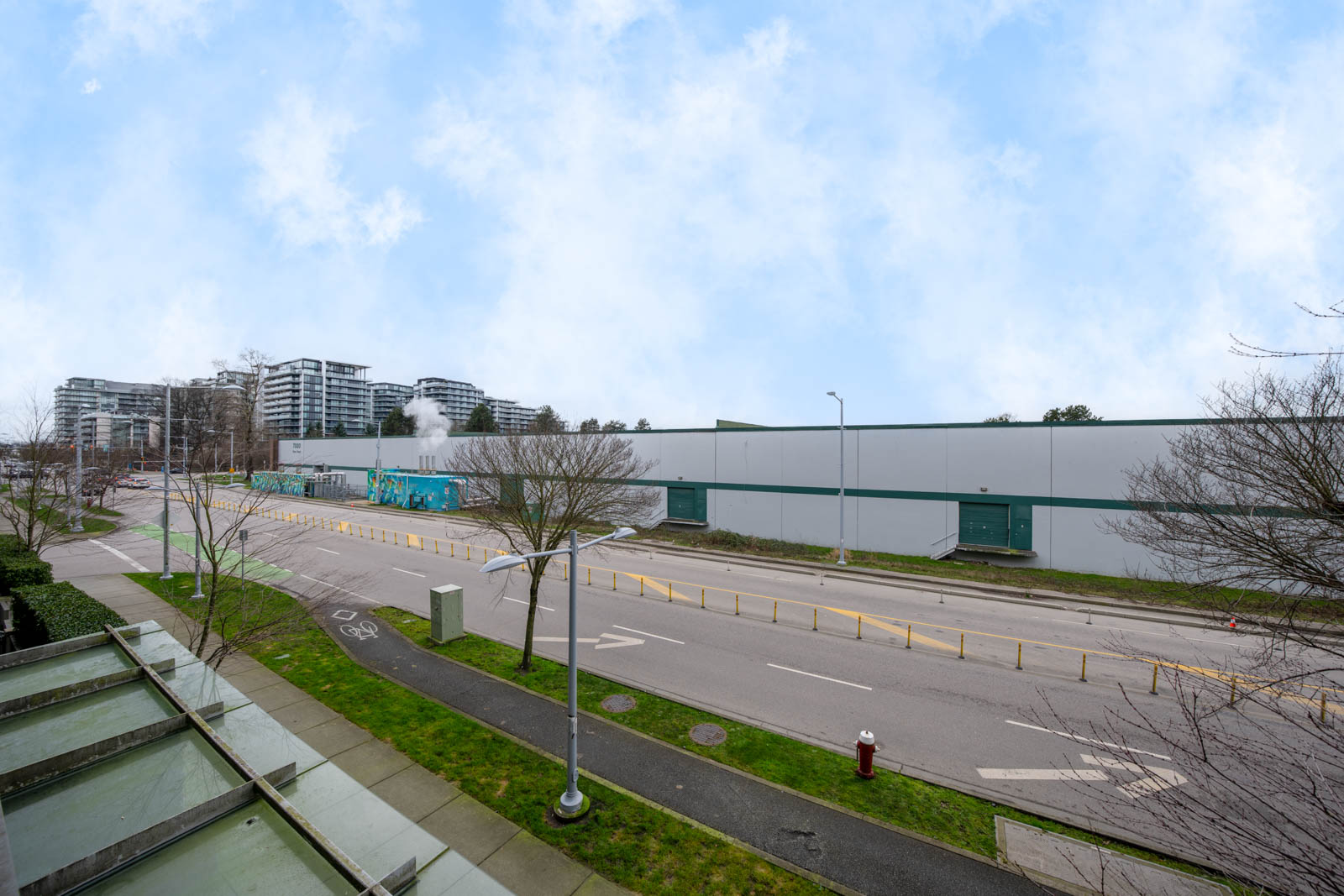 A wide street runs alongside a large, plain industrial building with some trees, bike lanes, and distant apartment buildings under a partly cloudy sky.