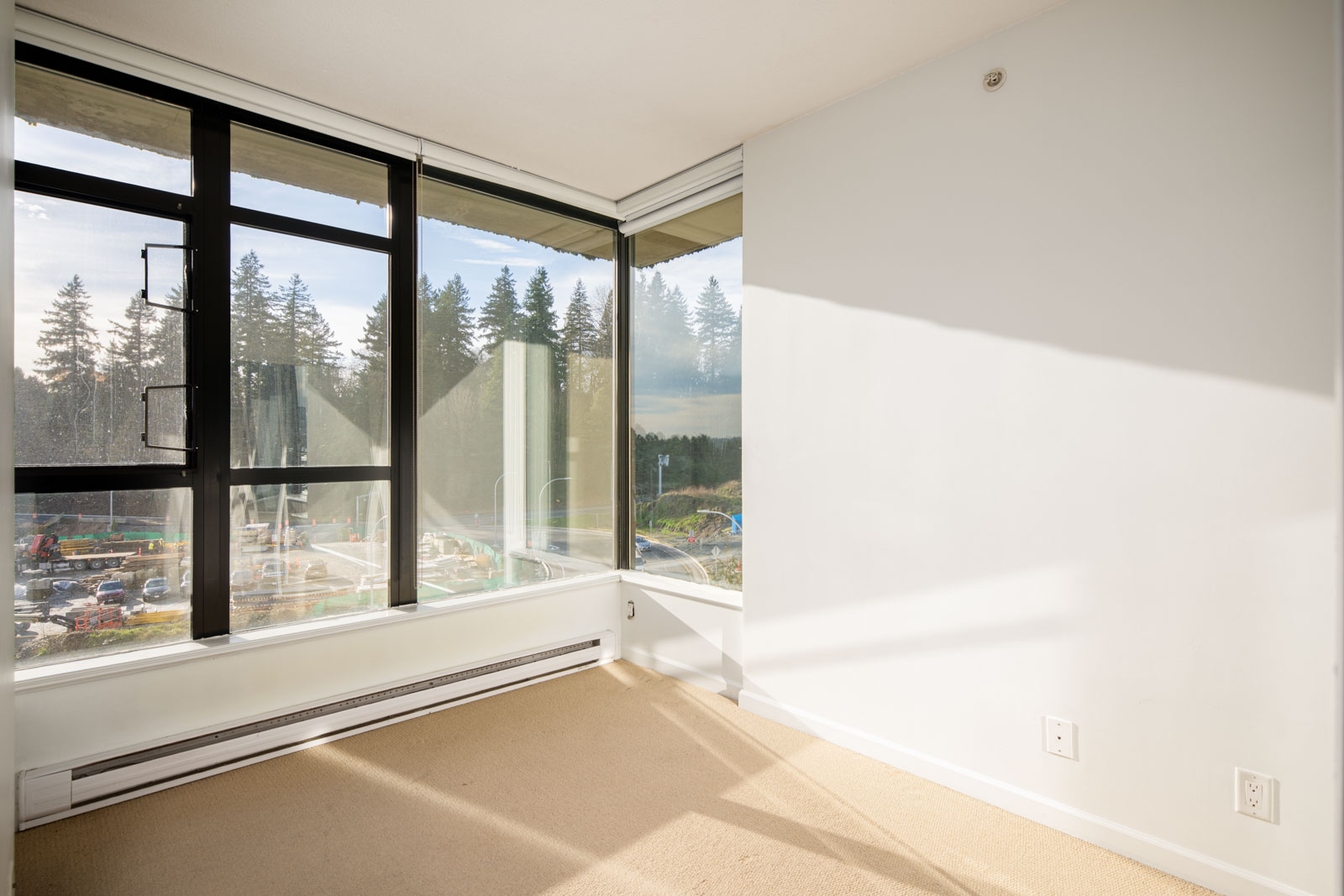 A small, empty room with beige carpet, white walls, and large corner windows overlooking trees and a parking lot on a sunny day.