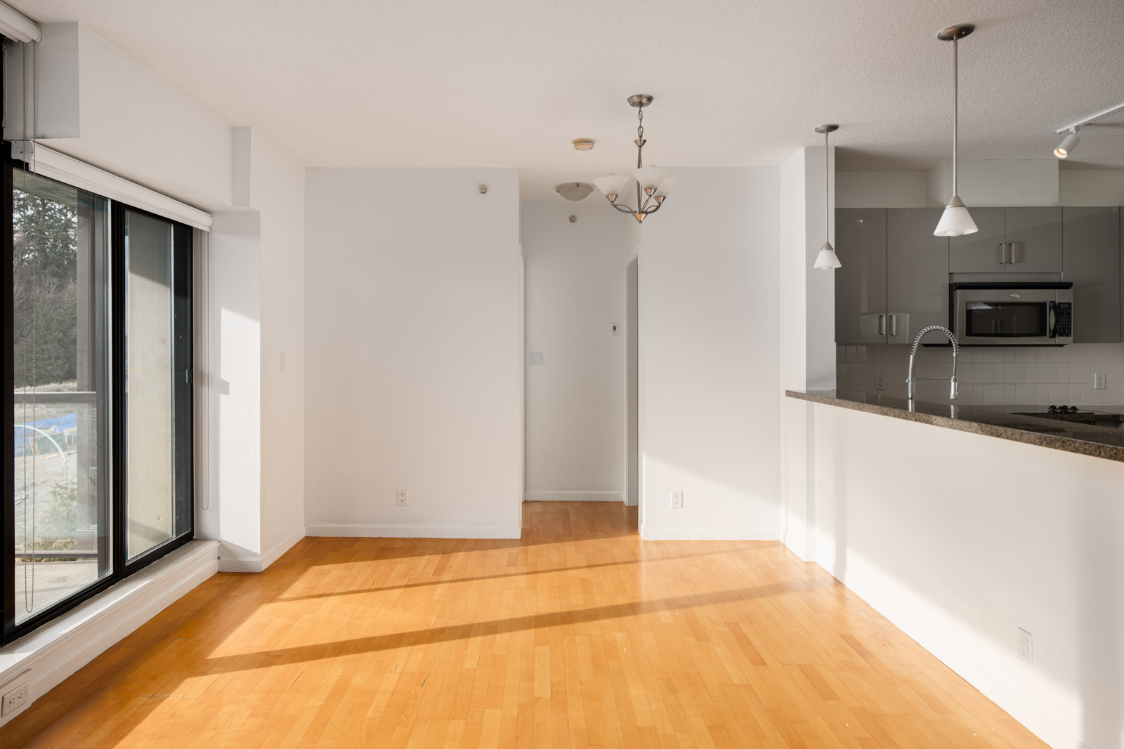 Empty, sunlit apartment interior with hardwood floors, white walls, large window, kitchen with grey cabinets, granite countertop, and pendant lights.