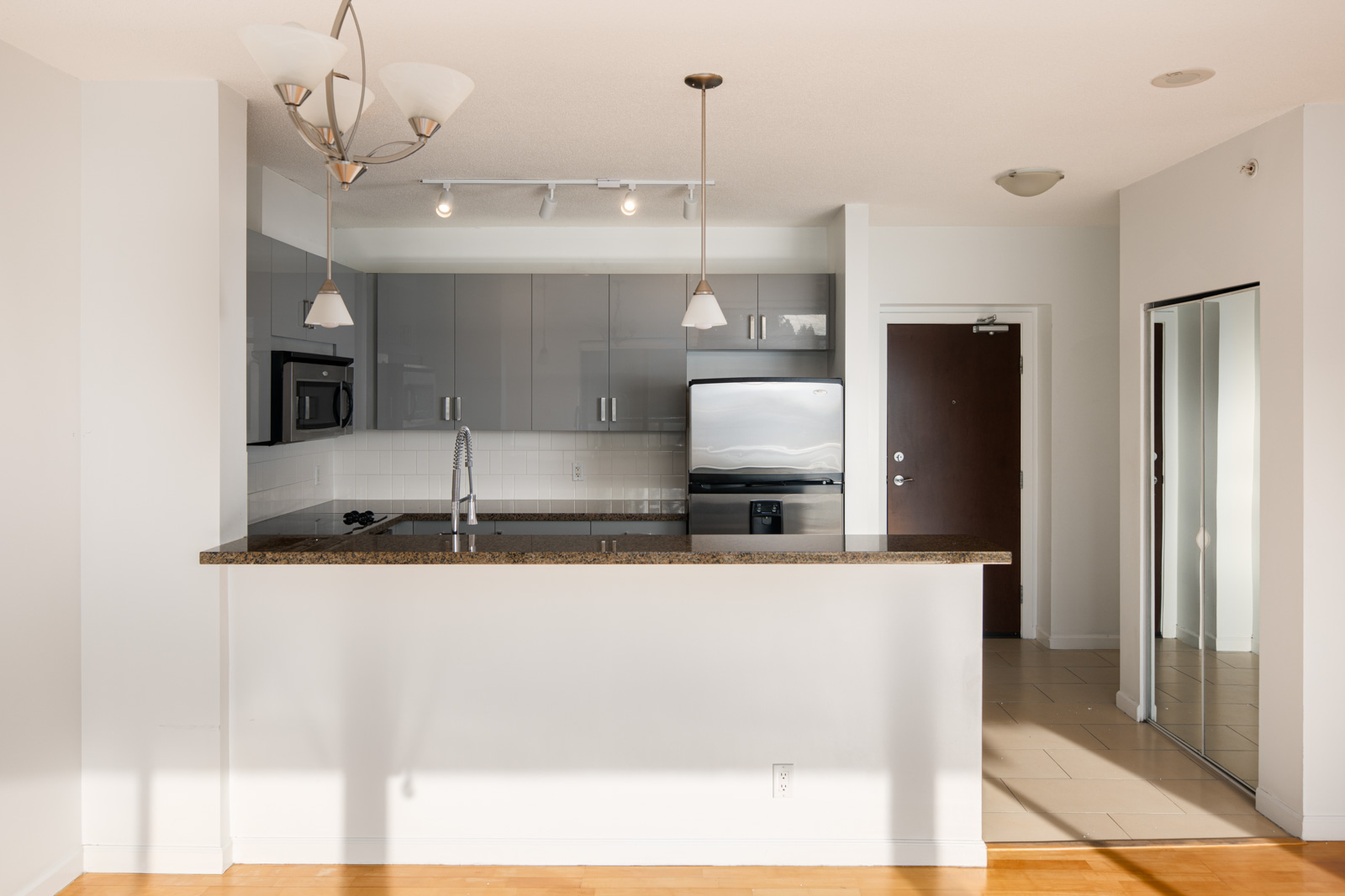 Modern kitchen with gray cabinets, stainless steel appliances, a granite countertop, pendant lights, and a dark brown entry door in the background.