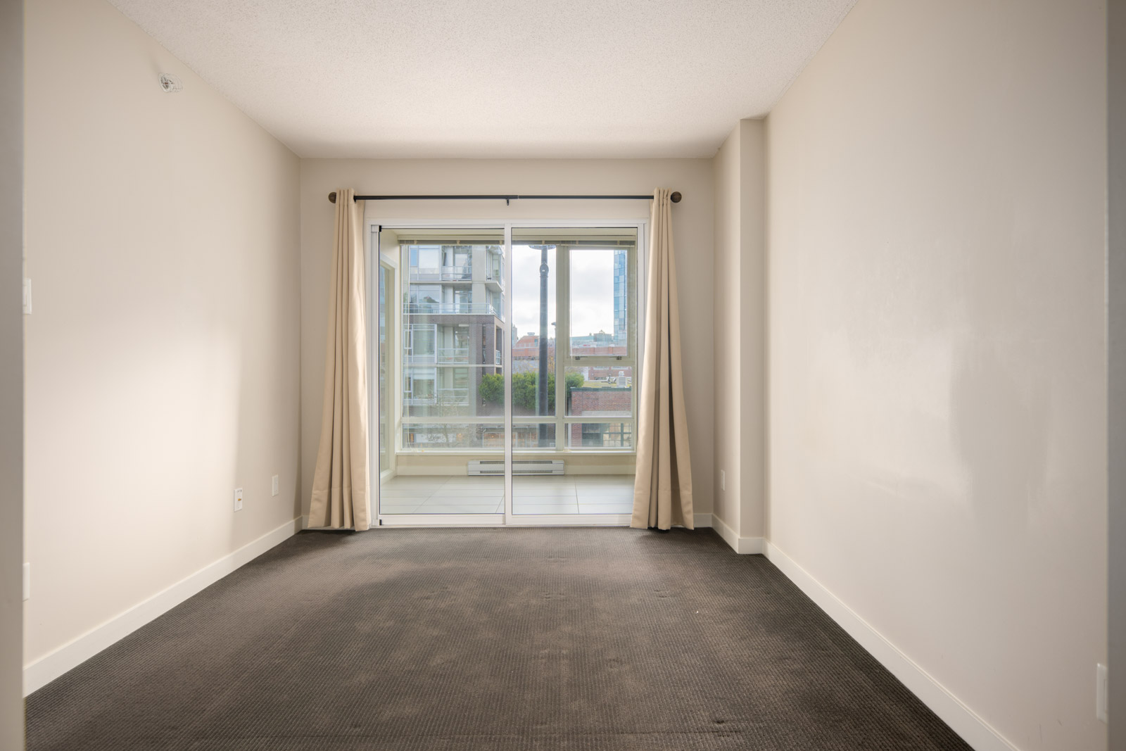 Empty, unfurnished room with beige walls, dark carpet, and sliding glass doors leading to a balcony with an urban view. Cream-colored curtains frame the window.